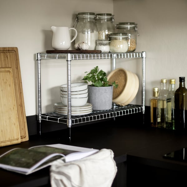 Kitchen counter with OMAR chrome shelf holding jars, bowls, and a plant. Wooden cutting board and bottles beside it.