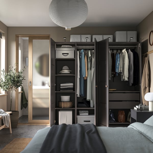 Modern bedroom with dark grey PAX wardrobe, hanging clothes, shelves, and a large mirror. Light blue bed, white pendant lamp, and green plant visible.