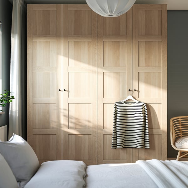 Natural wood PAX wardrobe, tall and narrow beside white bed, hanging shirt, large overhead light.