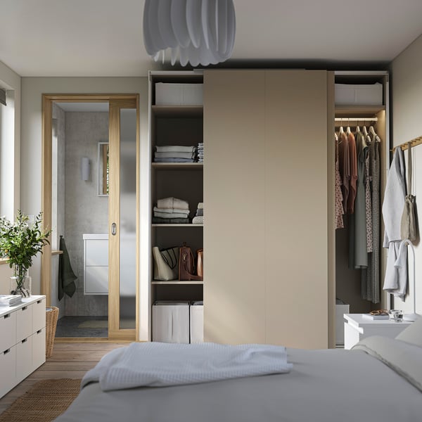 Modern bedroom with beige PAX wardrobe, open shelves, and neat clothes. White bed, pendant lamp, and bathroom entrance visible.