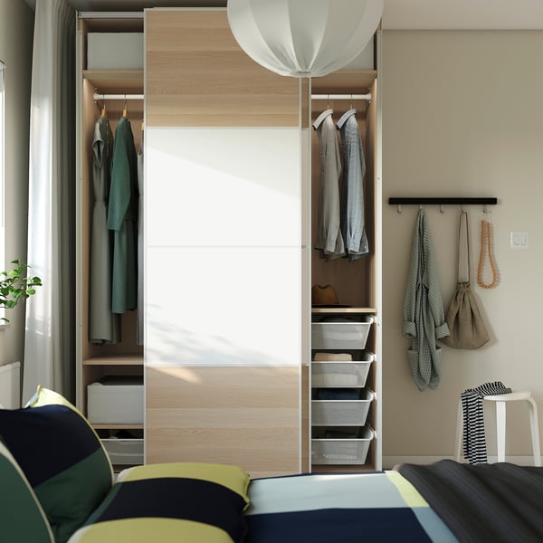 Modern bedroom with open PAX wardrobe displaying clothes, shelves, and organisers. Soft-closing doors, adjustable feet, and wall-mountable.