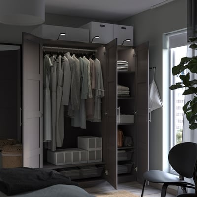 An open dark-colored PAX wardrobe in a bedroom, displaying clothes and storage boxes, lit by built-in lights.