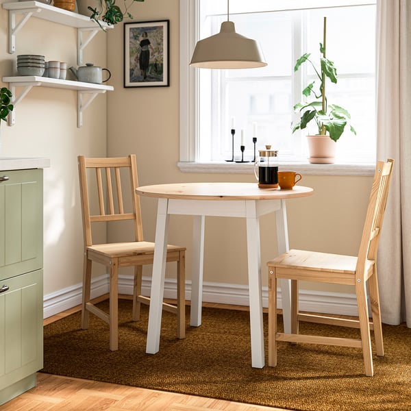 Cosy kitchen nook with small, white PINNTORP table and two matching chairs near a window, bright and inviting with rug and plants.