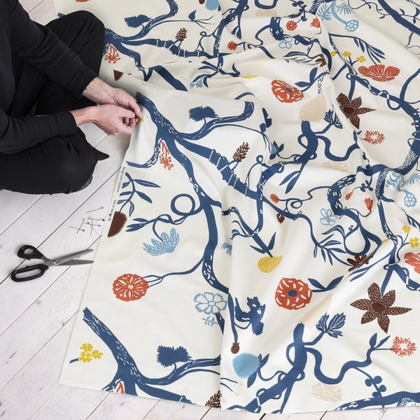 Person holding patterned fabric, scissors nearby. The fabric has blue branches, colourful flowers, and is laid out on the floor.