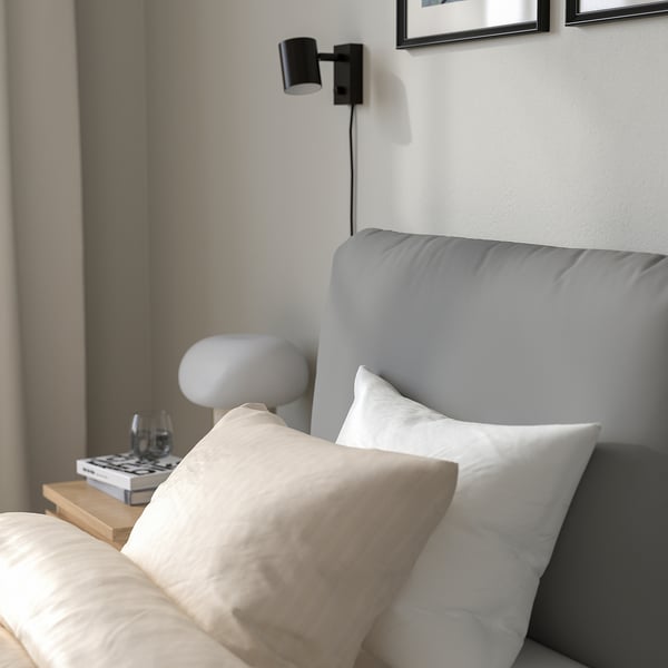 Modern bed with soft headboard; wall-mounted light, side table with book and glass.