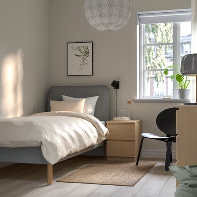 Modern bedroom: grey bed, white linens, natural light, bedside table, lamp, plant, black chair, rug.