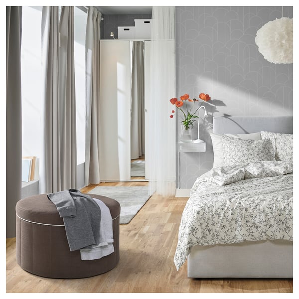 Modern bedroom: light floors, floral bed, footstool, large windows with grey curtains.
