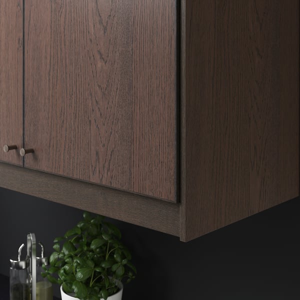 Wooden kitchen cabinet with simple knobs, displaying dark wood grain texture. Below, a potted plant is visible.