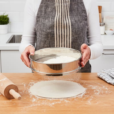 A person is baking, using a flour sifter over a mixing bowl, flouring the countertop with a rolling pin nearby.