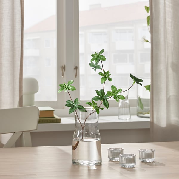 Clear glass vase with green plants on table, two small glass candles beside it. Behind window with sheer curtains.