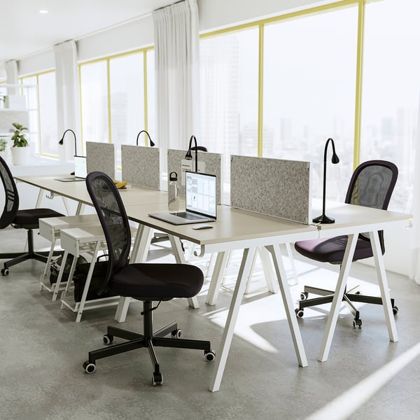 Modern office space with trottten white desks and black chairs, featuring large windows and natural light.