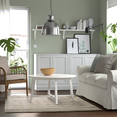 Contemporary living room with green walls, white furniture, and a wooden coffee table. Grey rug, hanging lamp, bookshelves, and balcony greenery.