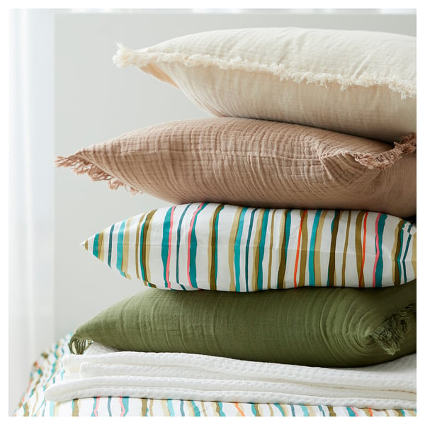 Four stacked pillows, top to bottom: white crinkled, beige fringed, striped multicoloured, and green solid with fringed edges.