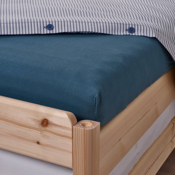 A bed with a blue cotton sheet tucked on a wooden frame. The sheet has elastic edges providing a snug fit.