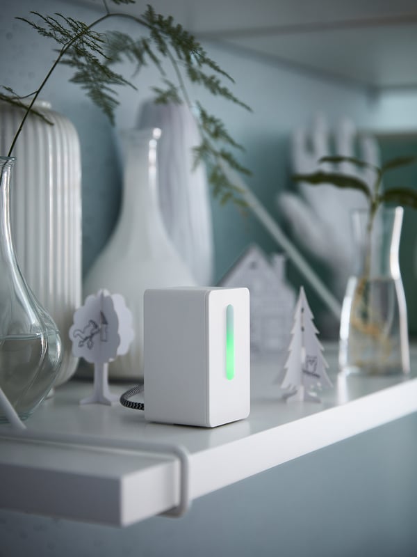 Small white device glows green on shelf with vases, decorative trees, and plants.