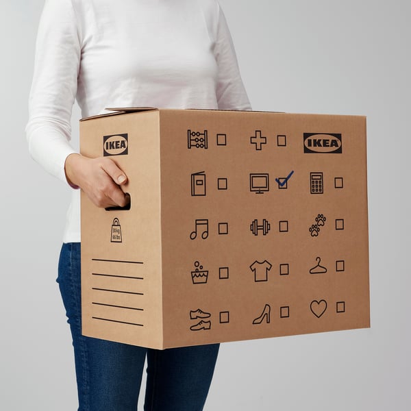 Oseba, ki drži trdno kartonsko škatlo ikea z izrezanimi ročaji za enostavno prenašanje. Škatla ima simbole in črte za označevanje vsebine.