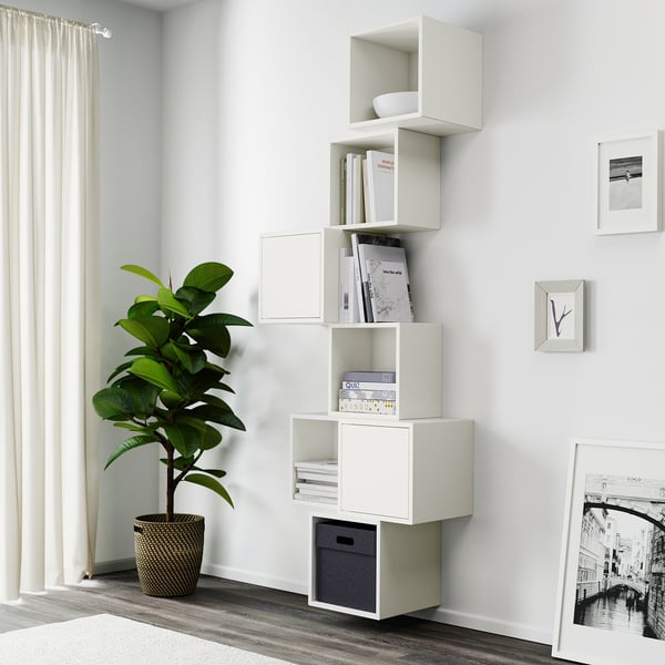 Bele modularne stenske police organizirajo knjige in dekor v bližini okna, ob njih pa je visoka zelena rastlina.