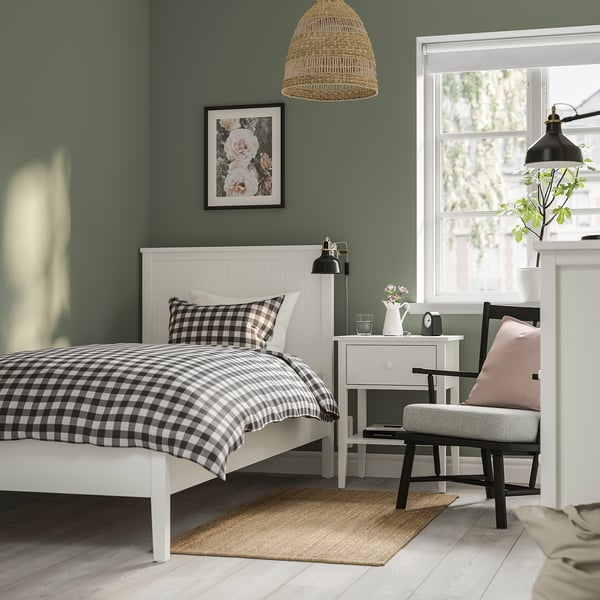 Karirasta črno-bela postelja ob zeleni steni, leseno vzglavje, nočna omarica s cvetjem, roza blazina na sivem stolu.
