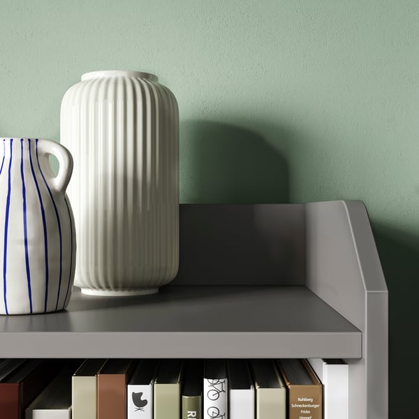 Dve vazi na sivi polici ob zeleni steni. Ena bela, rebrasta vaza in ena bela vaza z modrimi črtami. Sodoben, minimalističen dizajn.