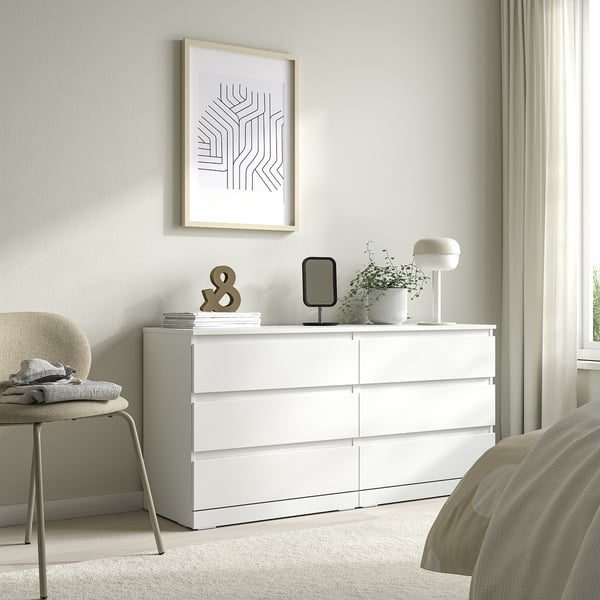 Bela LILLBERGET skrinja s šestimi predali in umetniškim tiskom zgoraj, stol levo, svetilka, rastlina na vrhu.