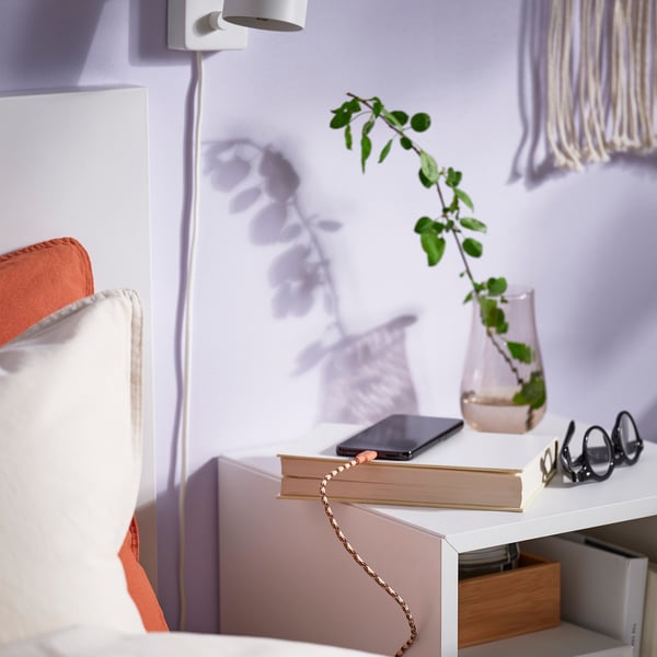 Udoben kotiček spalnice z belo posteljo, oranžno blazino, LILLHULT USB kabel za polnjenje telefona na knjigo in vazo z zeleno rastlino.