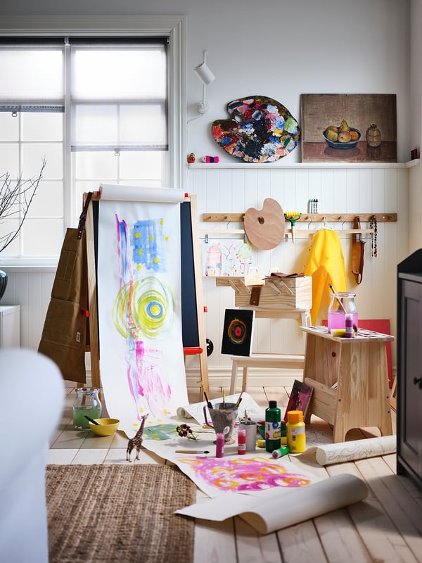 Umetniški studio s stojali, barvami, papirji in uokvirjenimi slikami.