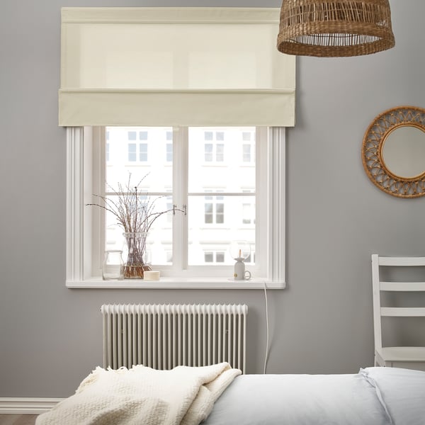 Bež RINGBLOMMA žaluzija na oknu, ki osvetljuje udobno spalnico z belo posteljo, pleteno odejo in ogledalom iz ratana na lestvi knjižne police.