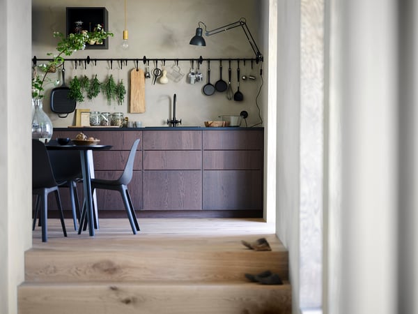 Sodobna kuhinja s temnimi lesenimi omarami, visečimi pripomočki in črno jedilno mizo s sivimi stoli.