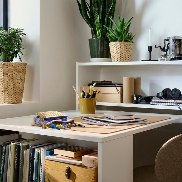 White desk with yellow cup of pens, notebooks, books, and plants. Small potted plants on shelves, drawer, compartments.