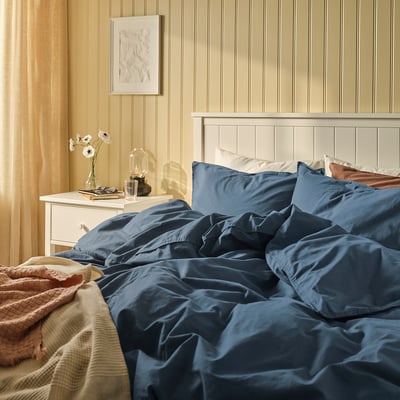 Bedroom with blue ÄNGSLILJA duvet cover on white bed, bedside table with flowers, on beige walls.