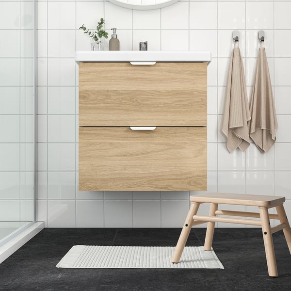 Minimalist bathroom with ALSTERN white bath mat, light wood vanity, bench, beige towels. Mat fits perfectly in front of vanity.