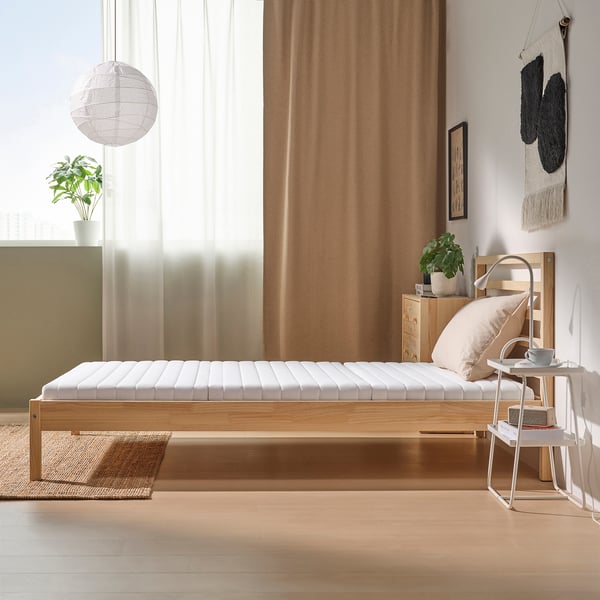 Minimalist bedroom with white bed, wooden frame, and large window. Decor includes plant, light, and tapestry.