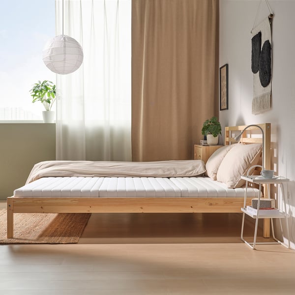 Cosy bedroom with beige mattress, wooden furniture, side table lamp, plant, large windows, and hanging textile.