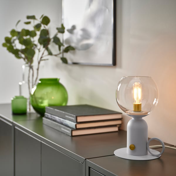 A modern grey ÅSKMULLER table lamp with a clear glass shade, sitting on a shelf next to a stack of books and green vases, casting a warm glow.