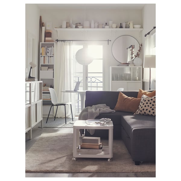 Bright living room with grey sofa, white coffee table, bookshelf, large window with white curtains, and various decorative items.