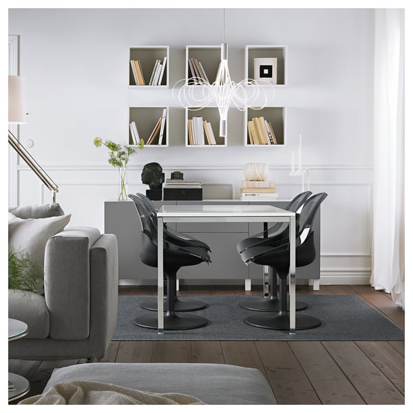 Modern dining area with glass-top table, BALTSAR chairs, and decor. Bright lighting and book-filled shelves enhance elegance.