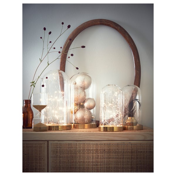 BEGÅVNING glass dome display with gold base, featuring string lights and decorative items inside.