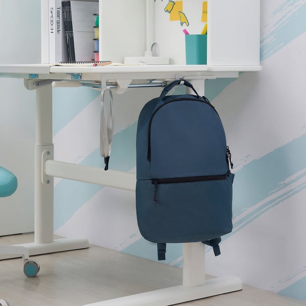 Blue backpack hangs on white desk with adjustable height and tiltable surface.