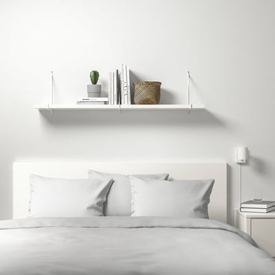 Minimalist bedroom with white BERGSHULT shelf above bed, holding plant, books, and basket. Bed has white pillows and headboard.