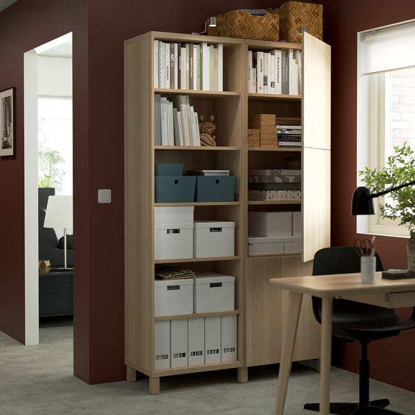 A wooden bookshelf with adjustable shelves storing books, boxes, and decorative items. It is positioned in a home office setting near a desk.