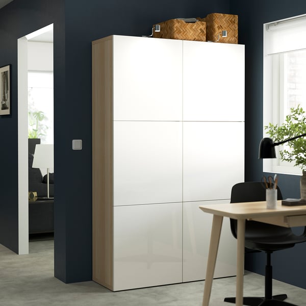 A white BESTÅ storage cabinet. It stands tall amongst a room with a desk and chair, highlighting its size and modern design.