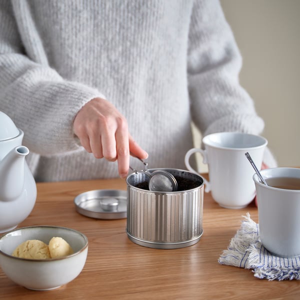 Person pours tea into silver BETTHAJ tin on wooden table with mugs and cookies.