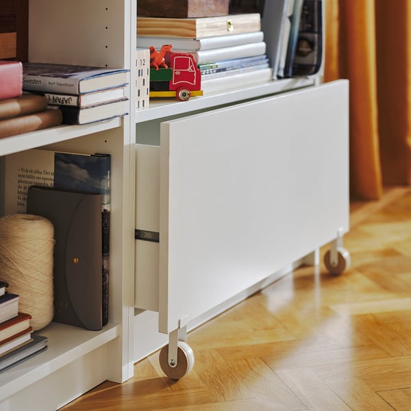 White BILLY bookshelf with drawers, wooden wheels, holding books, toy car, wooden floor.