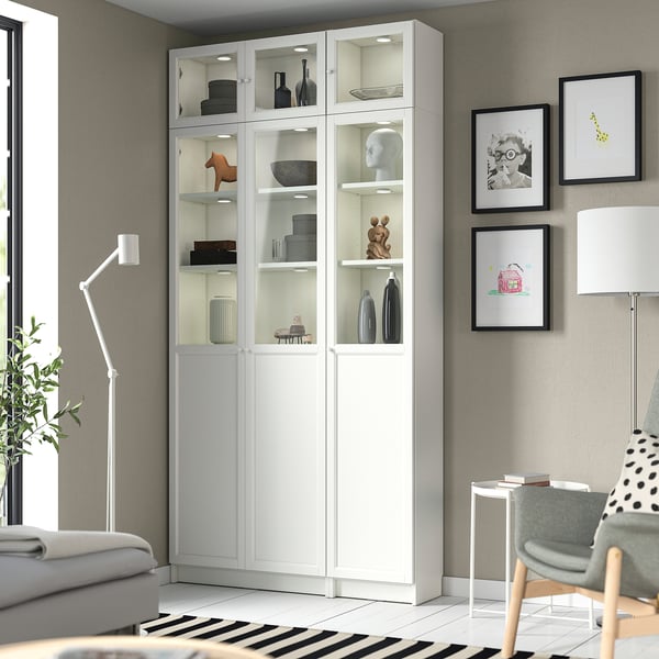 White BILLY bookcase with glass doors, displaying decor. Set in grey living room with sofa, chair, and wall art.