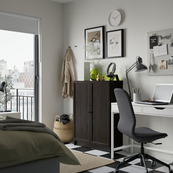Modern home office with BILLY bookcase in dark brown. Desk with chair, laptop, and decor against light grey wall. Cosy bedding in foreground.