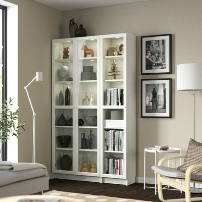 White BILLY bookcase with glass doors, filled with books and decorative items, tall and sleek, fits in living room with sofa and chair.