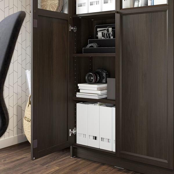 Open BILLY bookcase with adjustable shelves, displaying items and hiding clutter. Dark wood finish with glass doors showing organised storage.