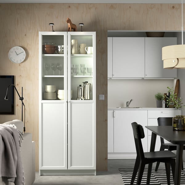 Modern kitchen with white BILLY cabinet, glass doors, and black dining table. Wooden walls, clock, and potted plants.