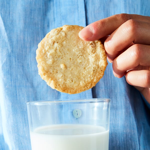 A hand holds a round oat biscuit above a glass of milk. Blue denim shirt background.