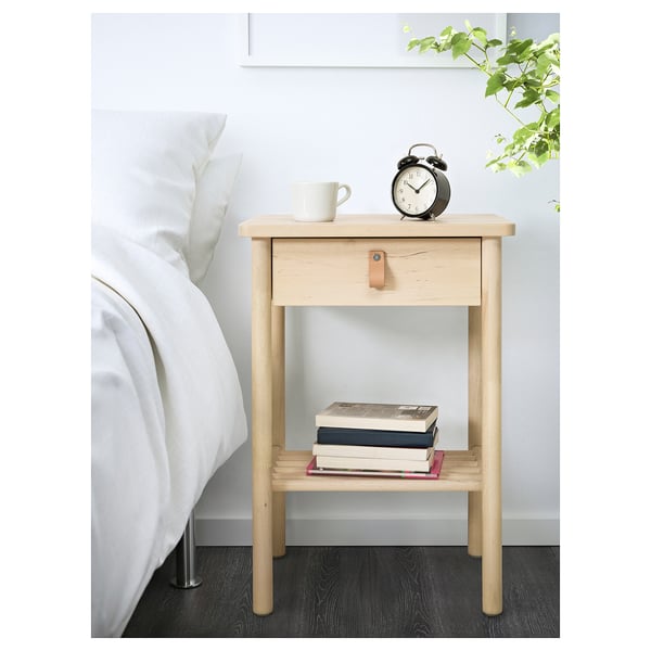 A light wooden bedside table with a drawer and lower shelf. It holds a stack of books and a clock.